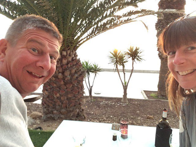 Steve who has MPN, sits smiling with his wife at a table, in front of a palm tree.