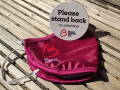 A Blood Cancer UK facemask placed on a table with a Blood Cancer UK please stand back I'm shielding badge, placed on top of it.