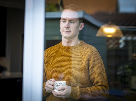 Rob, who lives with CML, stands looking out of a window, holding a mug.