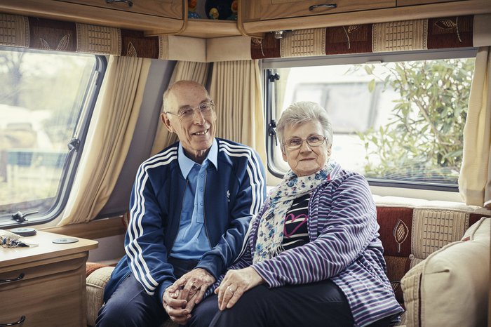 Trevor, who has chronic lymphocytic leukaemia (CLL), sits smiling, holding hands with his wife on the sofa at the back of a caravan.