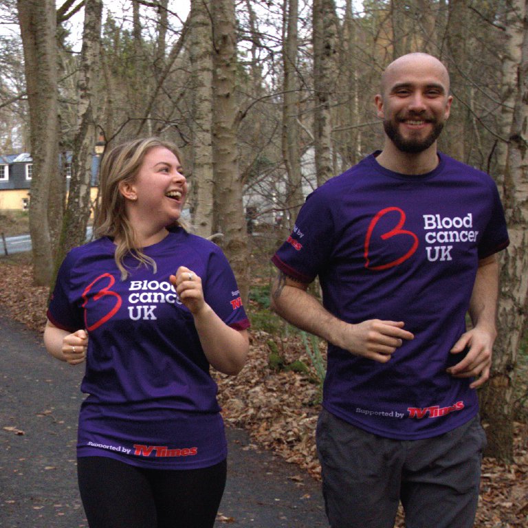 Two runners - a man and a woman - enjoy a run down a forest path. They both wear purple Blood Cancer UK t-shirts.