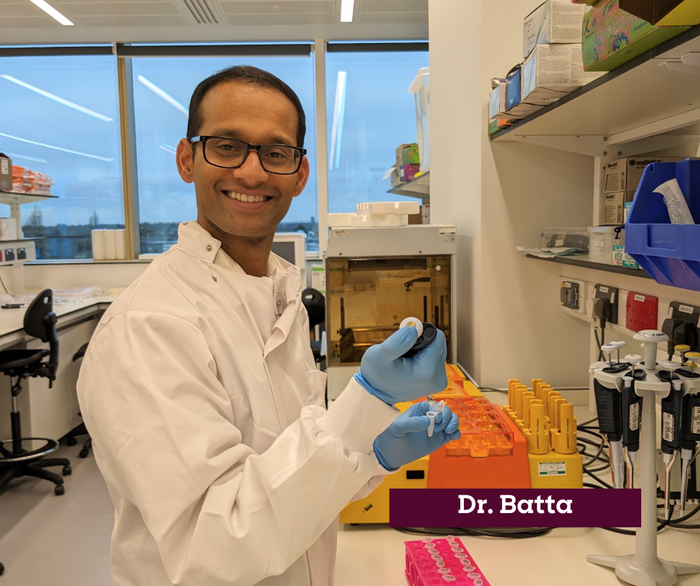 Dr Kiran Batta working in the lab smiling, wearing a white lab coat.
