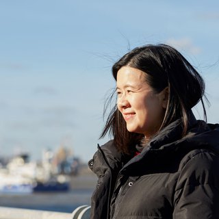 Jacqueline, in remission from DLBCL, stands smiling in the sunshine with a city skyline blurred in the background.