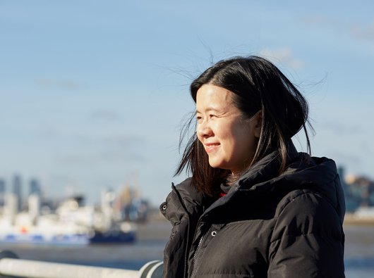 Jacqueline, in remission from DLBCL, stands smiling in the sunshine with a city skyline blurred in the background.