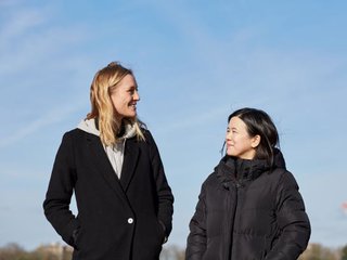 Jacqueline, in remission from DLBCL, stands smiling with her friend on a clear sunny day.