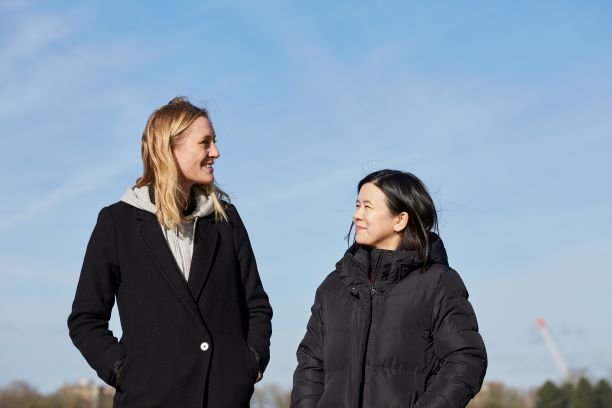 Jacqueline, in remission from DLBCL, stands smiling with her friend on a clear sunny day.