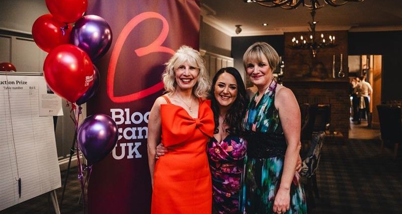 A group of women at a Let's Beat It Blood Cancer UK event, standing in smart clothing next to banners and balloons.