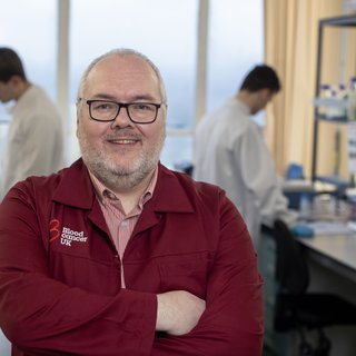 Professor Tonks stood in the lab, with his arms folded, wearing a red Blood Cancer UK lab coat.