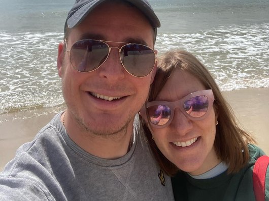 Andy and his wife are standing on the beach, with their back to the sea. They are wearing sunglasses, and smiling at the camera.