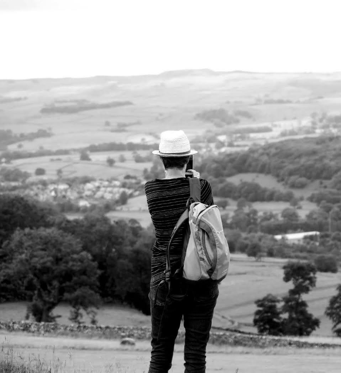 Black and white photo of Becky. Becky is in a field, seen from behind. She's taking a photo on her phone.