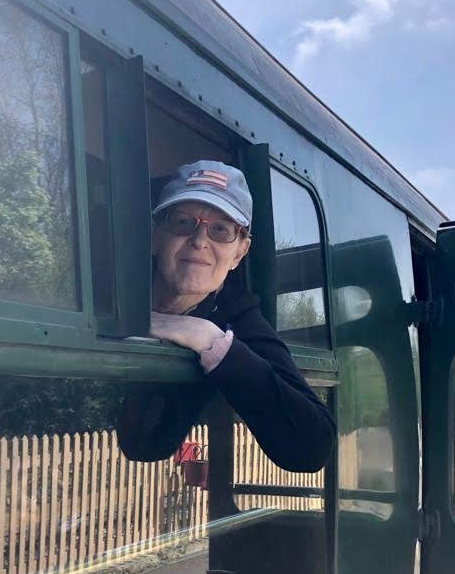 Becky, wearing a cap and glasses, looking out of an old fashioned train carriage window.