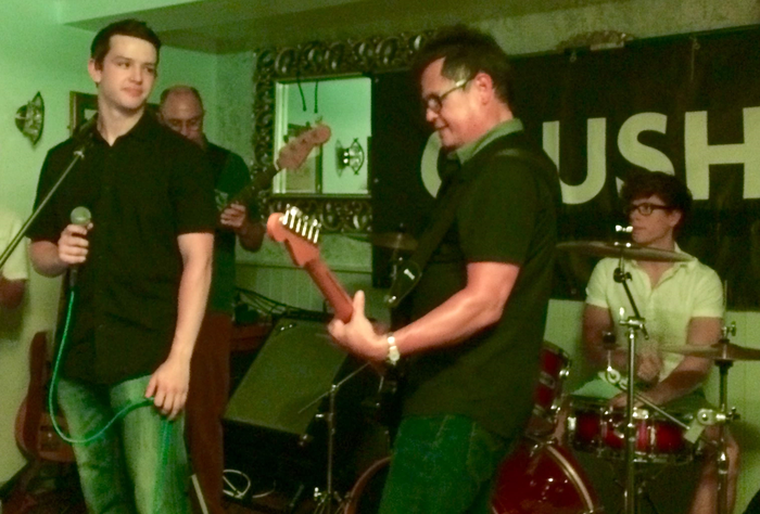 Ben and his father (who has AML) stand on stage with a band, Ben is holding a microphone whilst his dad plays electric guitar.