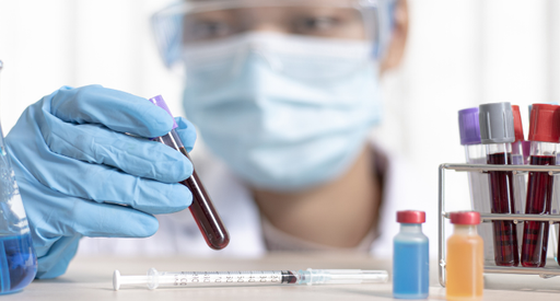 A researcher wearing a facemask inspects a vial of blood on a table with other vials.