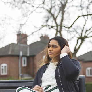 A young woman with blood cancer sits on a bench looking pensive, in front of a row of houses.