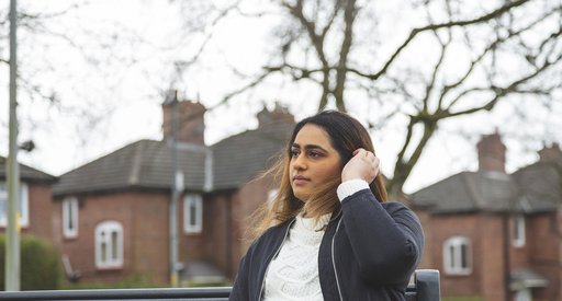 A young woman with blood cancer sits on a bench looking pensive, in front of a row of houses.