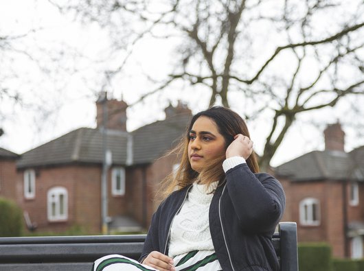 A young woman with blood cancer sits on a bench looking pensive, in front of a row of houses.