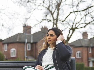 A young woman with blood cancer sits on a bench looking pensive, in front of a row of houses.