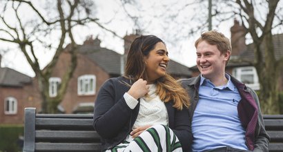 A young couple sit laughing together on a bench, in front of some houses.