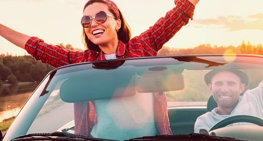 A very happy woman stands up in a convertible car with her hands aloft.