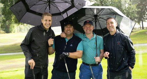 Four men stand under golf umbrellas during a brief rain shower and smile for the camera