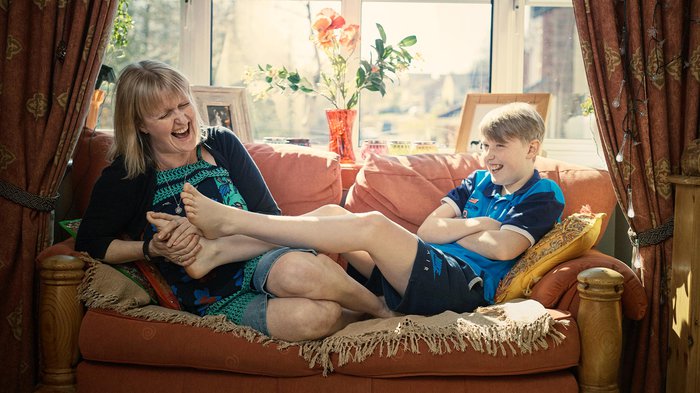 A mother and child laughing together on their sofa at home, the son's feet on his mother's lap.