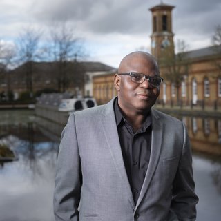 David, wearing a grey suit jacket and black shirt which is open at the neck. Behind David there is a canal boat and building.
