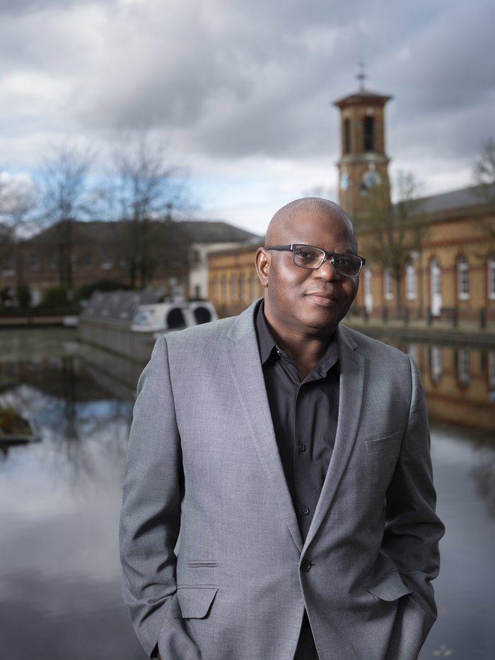 David, wearing a grey suit jacket and black shirt which is open at the neck. Behind David there is a canal boat and building.