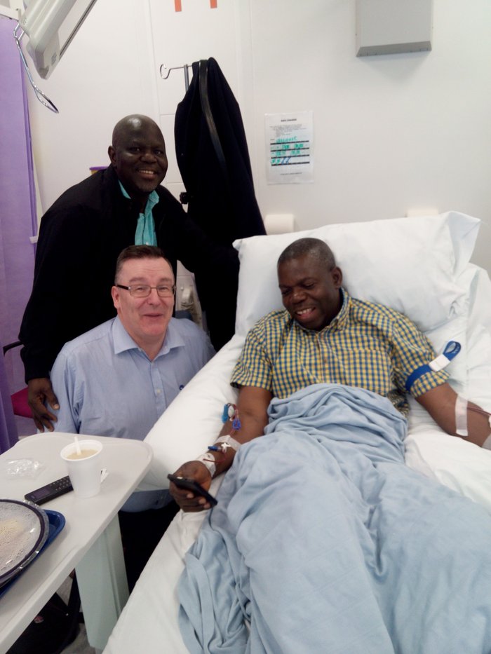 David's brother in hospital donating his stem cells. David's brother is smiling while lying in a hospital bed, with a thin tube in each arm. David and another man are next to the bed, smiling at the camera.