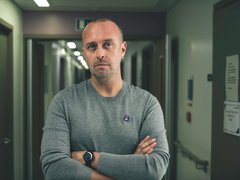 A vignette of a man folding his arms and looking seriously into the camera. In the background there is a dimly lit hospital corridor.