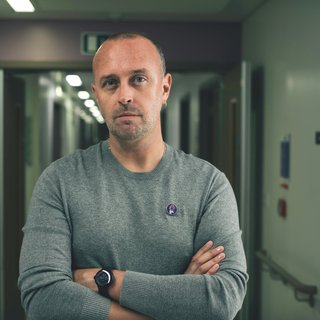 A vignette of a man folding his arms and looking seriously into the camera. In the background there is a dimly lit hospital corridor.