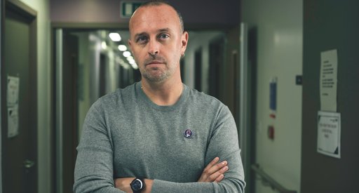 A vignette of a man folding his arms and looking seriously into the camera. In the background there is a dimly lit hospital corridor.