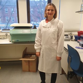 A women stood in the lab smiling wearing a white lab coat.