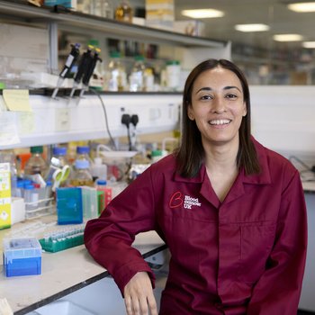 Dr Jasmeen Oberoi stood smiling in the lab wearing a Blood Cancer UK lab coat.