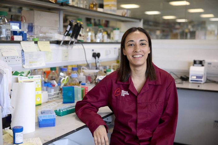 Dr Jasmeen Oberoi stood smiling in the lab wearing a Blood Cancer UK lab coat.