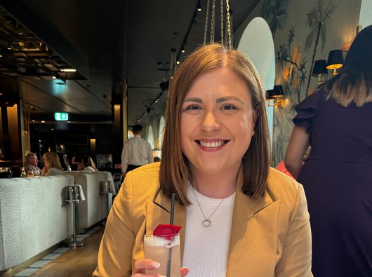 A young woman (Emily) sitting at a restaurant table. She's smiling and holding a drink.