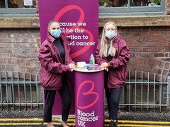 Two Blood Cancer UK members of staff, wearing masks, smiling at a bucket collection, in front of Blood Cancer UK merchandise.
