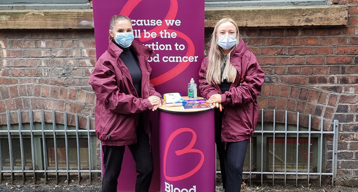Two Blood Cancer UK members of staff, wearing masks, smiling at a bucket collection, in front of Blood Cancer UK merchandise.