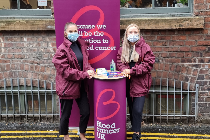 Two Blood Cancer UK members of staff, wearing masks, smiling at a bucket collection, in front of Blood Cancer UK merchandise.