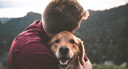 Man in burgundy shirt cuddles golden retriever in wilderness landscape.