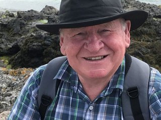 An older man - Gerald - in the countryside. He wears a brimmed hat, check shirt and has a backpack. The image is closely cropped with Gerald looking direct to camera and smiling.