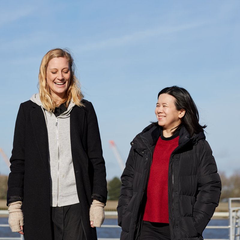 Two females standing side by side smiling in the sun with water in the background