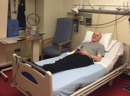 Graeme is lying on his back, on his hospital bed, with his head propped up on the pillow. He is looking at the camera and smiling.