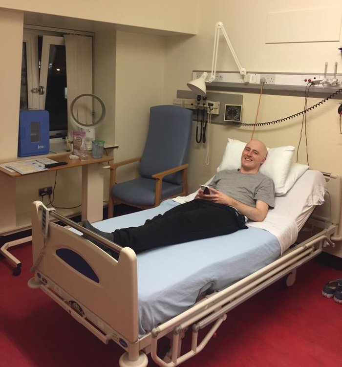 Graeme is lying on his back, on his hospital bed, with his head propped up on the pillow. He is looking at the camera and smiling.