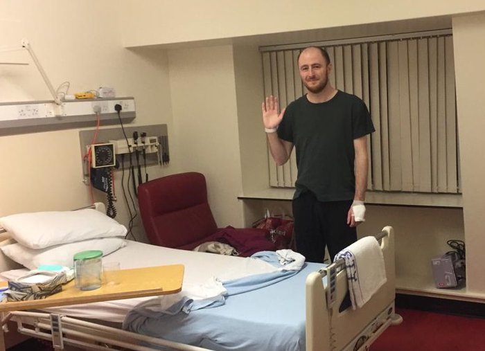 Photo of Graeme standing up behind his bed in his hospital room. He is smiling, and you can see a bandage around one hand, and a hospital wrist-band on the other hand.