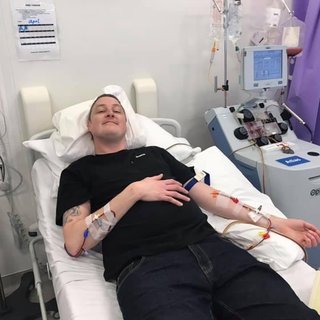 Grant having his T-cells collected. He's smiling while lying on a hospital bed. He's connected by thin tubes in each arm to an apheresis machine (a machine that separates blood) that's next to the bed.