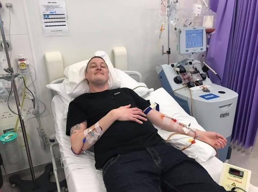 Grant having his T-cells collected. He's smiling while lying on a hospital bed. He's connected by thin tubes in each arm to an apheresis machine (a machine that separates blood) that's next to the bed.