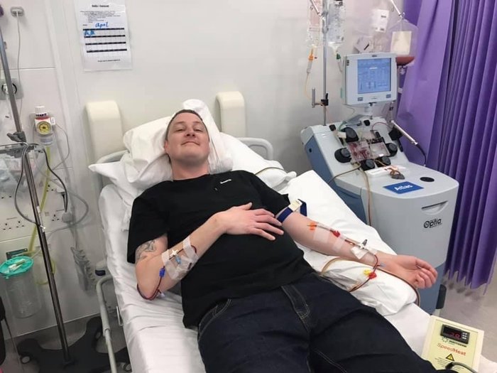 Grant having his T-cells collected. He's smiling while lying on a hospital bed. He's connected by thin tubes in each arm to an apheresis machine (a machine that separates blood) that's next to the bed.