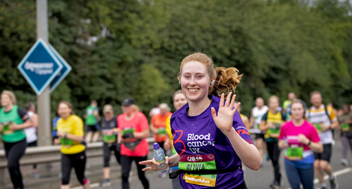 A woman in a purple Blood Cancer UK T shirt waves as she runs in a marathon.