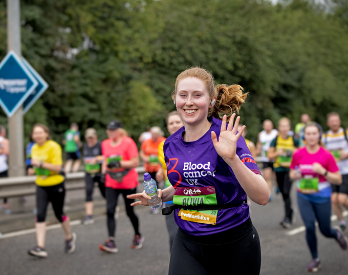 A woman in a purple Blood Cancer UK T shirt waves as she runs in a marathon.