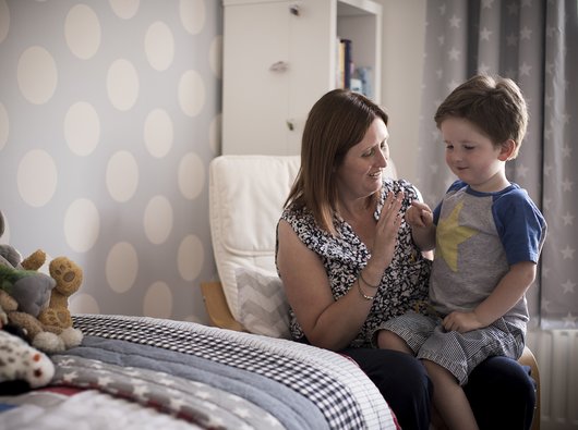 Hugo, in remission from leukaemia, propped on her knee and looking towards a grouping of cuddly toys.